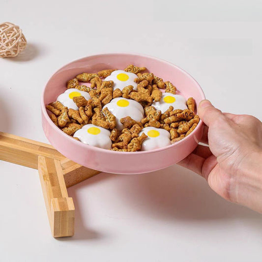 Pink bowl filled with dog food shaped like eggs and sausage, held by a hand on a light background.